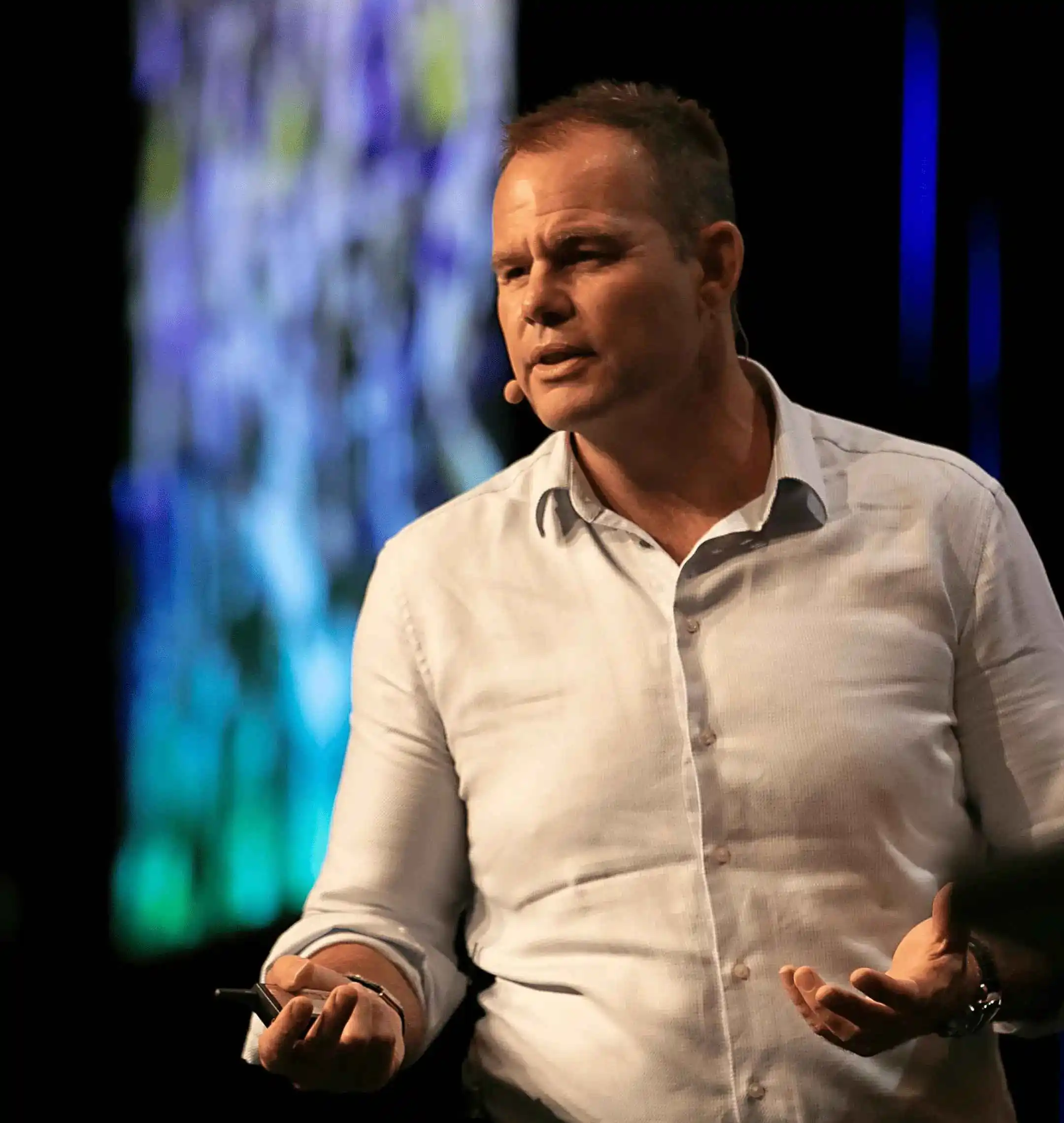 A man in a light-colored button-up shirt speaks on stage, gesturing with one hand and holding a pen in the othe